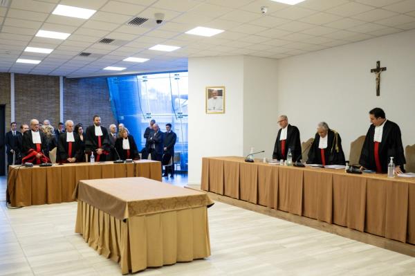 A room with three tables, one empty in the center and two lining the walls, with men in black robes standing behind them at the Vatican tribunal.