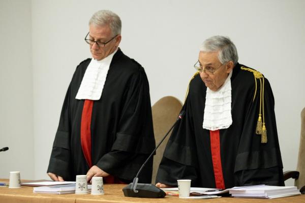 Two men in black robes with white ceremo<em></em>nial collars stand at a table fitted with micropho<em></em>nes to announce the verdict in the Vatican tribunal.