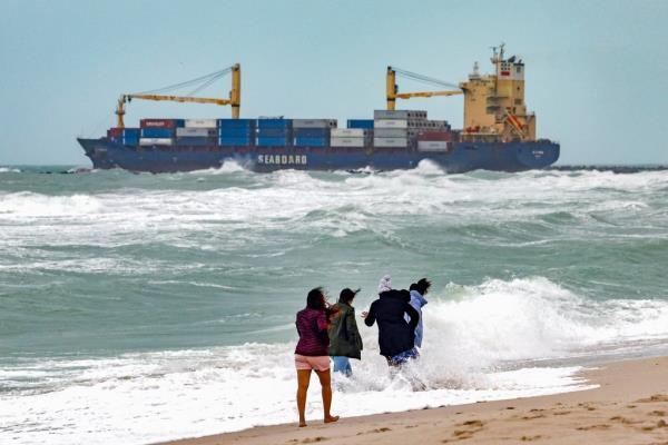 A group of people walking on a beach in Florida with choppy waves, as a co<em></em>ntainer ship sails in the water beyond them.